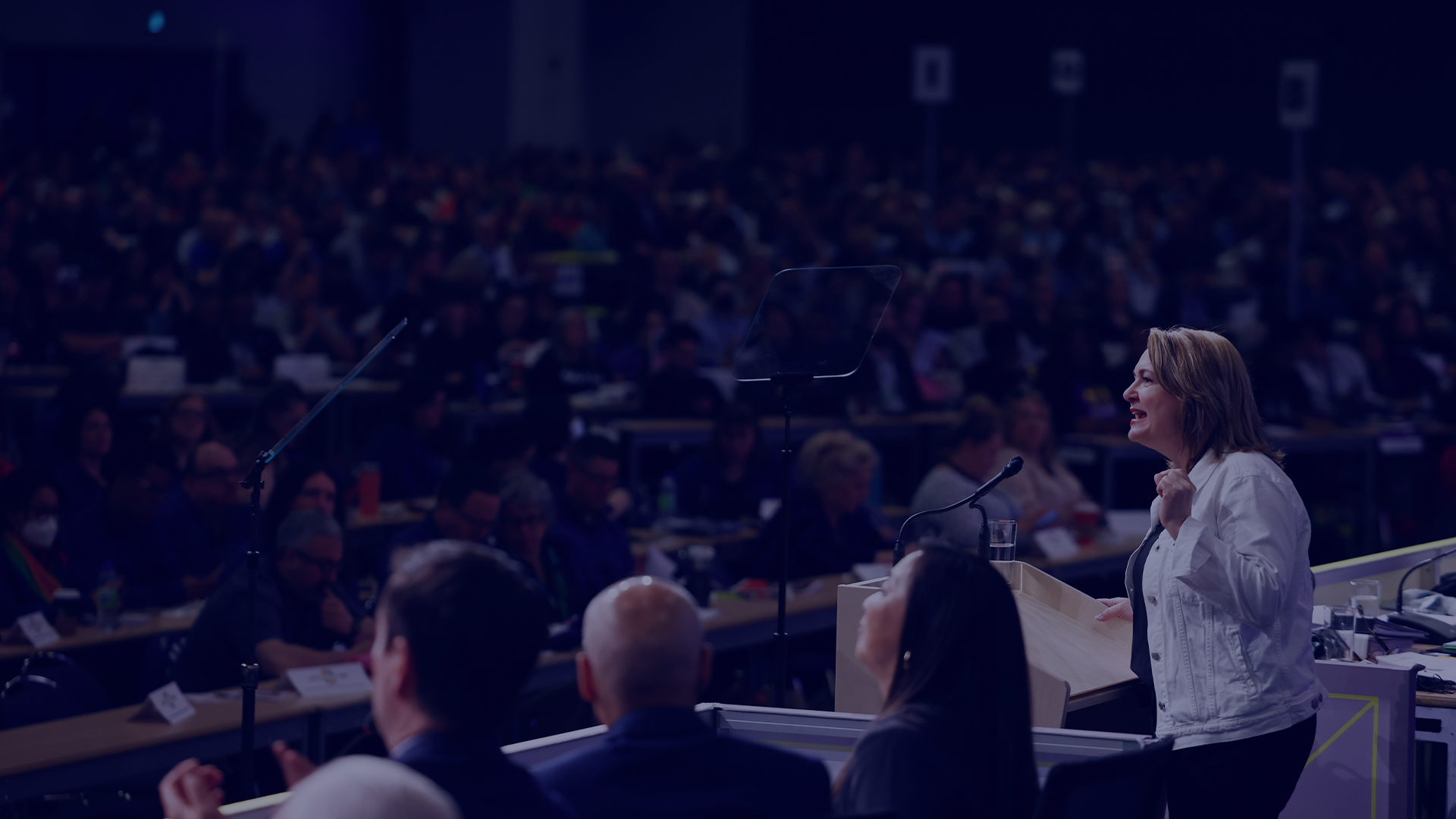 a female standing on a stage speaking to an audience of people
